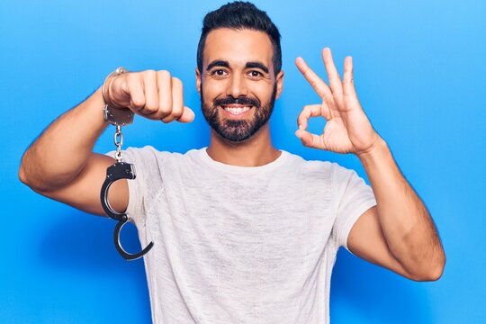 Young hispanic man holding prisoner handcuffs doing ok sign with fingers, smiling friendly gesturing excellent symbol