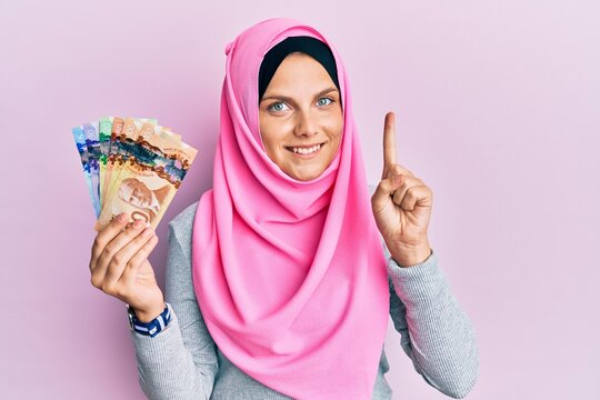 Young caucasian woman wearing islamic hijab holding canadian dollars banknotes smiling with an idea or question pointing finger with happy face, number one