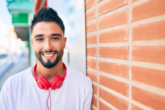 Young arab man smiling happy using headphones leaning on the wall at the city.
