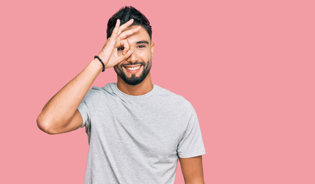 Young man with beard wearing casual grey tshirt doing ok gesture with hand smiling, eye looking through fingers with happy face.