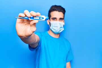 Young handsome man with beard wearing medical mask holding thermometer looking positive and happy standing and smiling with a confident smile showing teeth