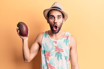 Young handsome man with beard wearing summer hat and t-shirt holding mango scared and amazed with...