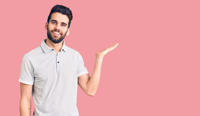 Young handsome man with beard wearing casual polo smiling cheerful presenting and pointing with palm of hand looking at the camera.