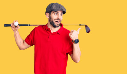 Young handsome man with beard playing golf holding club and ball pointing thumb up to the side...