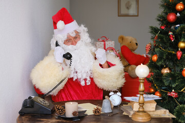 adult santa claus in red suit sits at the table, calls on old phone, tree is beautifully decorated with balls, the concept of christmas, new year celebration, holiday sales and discounts, sylvester