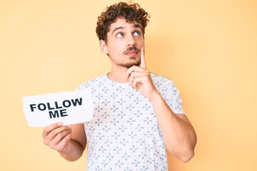 Young caucasian man with curly hair holding follow me message paper serious face thinking about...