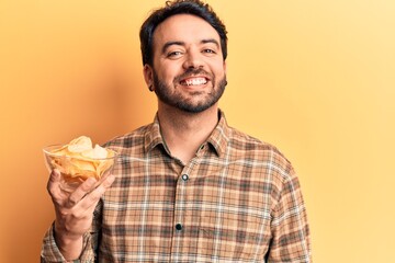 Young hispanic man holding potato chip looking positive and happy standing and smiling with a confident smile showing teeth