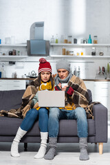 Fototapeta na wymiar cold couple in warm hats and gloves sitting on sofa under plaid blanket and watching movie on laptop