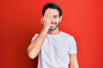 Young hispanic man wearing casual white tshirt covering one eye with hand, confident smile on face and surprise emotion.