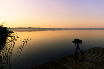Fototapeta na wymiar Photography spot with camera in the front by a colorful sunrise on a calm lake with reeds on the shore
