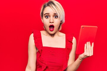 Young beautiful blonde woman holding touchpad scared and amazed with open mouth for surprise, disbelief face
