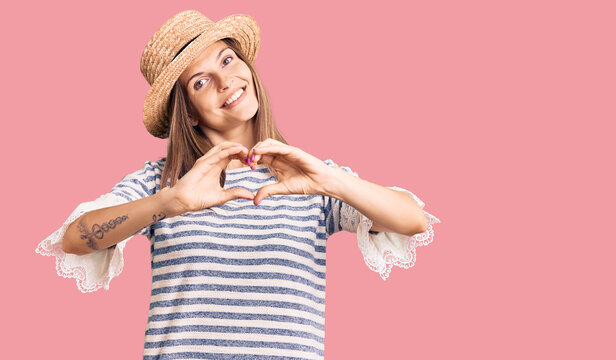 Beautiful caucasian woman wearing summer hat smiling in love doing heart symbol shape with hands. romantic concept.