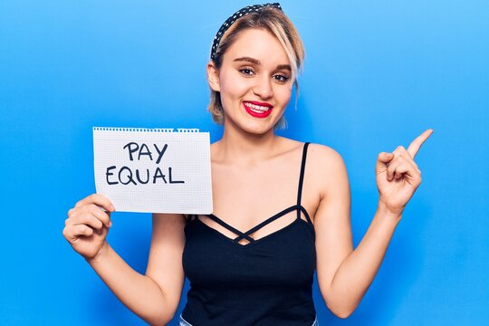 Young beautiful blonde woman holding pay equal paper smiling happy pointing with hand and finger to the side