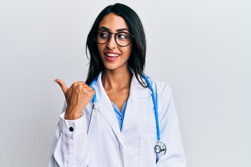 Beautiful hispanic woman wearing doctor uniform and stethoscope pointing thumb up to the side...