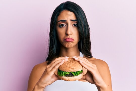 Beautiful Hispanic Woman Eating A Tasty Classic Burger Depressed And Worry For Distress, Crying Angry And Afraid. Sad Expression.