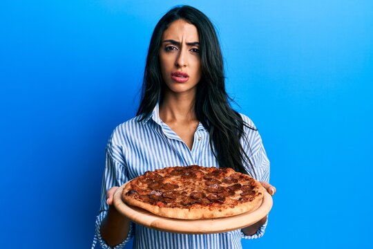 Beautiful hispanic woman holding tasty pepperoni pizza clueless and confused expression. doubt concept.