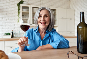 Happy 60s mature woman drinking wine video calling friend at home. Old middle aged lady holding glass talking to web cam by online social distance chat meeting sit at kitchen table. Webcam view.
