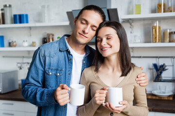 pleased couple standing in kitchen with closed eyes while holding cups of coffee
