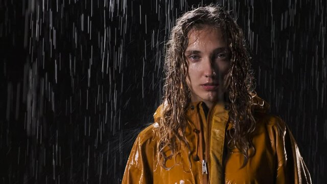 Portrait of woman who stands in the pouring rain at night on the street in yellow raincoat. Nice cute girl with wet hair on black background looks at the camera.
