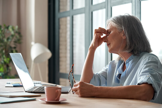 Tired old senior business woman taking off glasses suffers from eyestrain, stress, headache, fatigue after computer use work in office. Exhausted sick mature lady feels eye strain problem at workplace