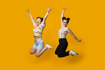 Fototapeta na wymiar Caucasian twins jumping and gesturing happiness on a yellow studio wall while wearing summer clothes and glasses
