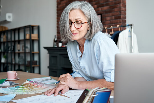 Middle aged stylish woman fashion designer drawing sketches in studio office. Mature old adult elegant grey-haired lady dressmaker small business owner creating new fashion design cloth in atelier.