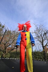 Halloween party outdoors.Scary  clown with red hair jumping in a trampoline on autumn trees and blue sky background. Creepy clown costume. Festival and carnival. 