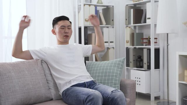 asina man stretching and leaning back into sofa, and putting hand behind head and crossing legs is bored.