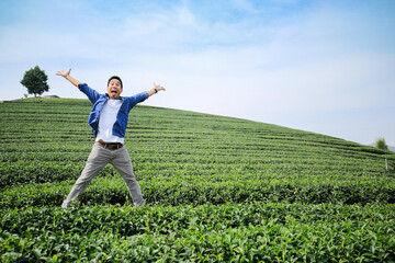 Fototapeta premium A happy man is jumping happily in tea plantation with hill and blue sky background. Wide angle shot.