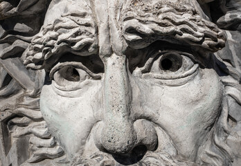 Close up of male sculpture made out of concrete. The men's face has wavy brows and a strong nose. Porous grunge concrete or rock texture from being exposed to the elements. Outdoor park water feature.