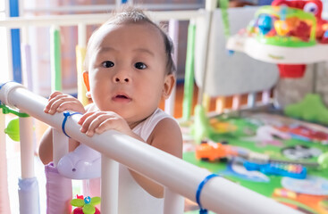 Cute little Asian baby boy toddler playing and looking up camera in the crib at home. Nursery for a young child. Breastfeeding or early childhood development concept. Copy space