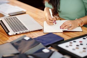 Fototapeta premium Close-up image of female atelier owner sitting at table with opened laptop and writing in planner