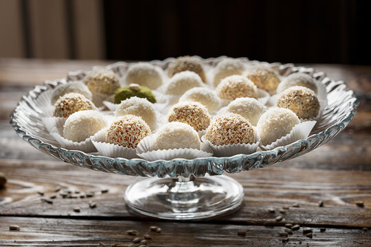 Glass cake stand full of variety of homemade handmade energy ball candies made of various nuts such as coconut, peanut, pistachio nuts on dark wooden table at kitchen. Horizontal orientation image