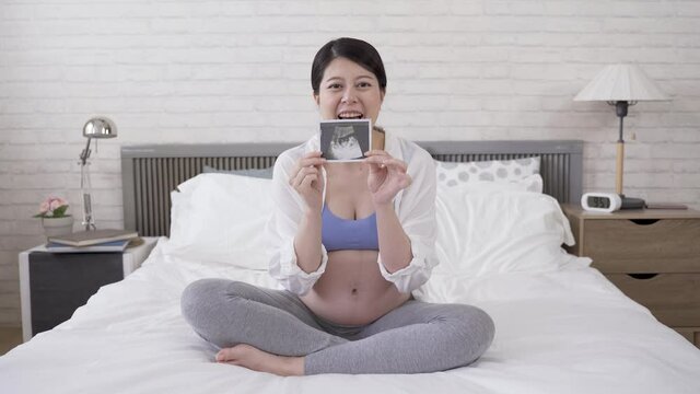 asian lady is shooting an online video in bedroom, waving hand and showing her latest ultrasound image to her friends
