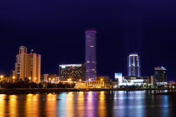 Cityscape with skyscrapers and other modern tower buildings with colorful illumination standing on the bank of river with reflections in it against dark blue sky at night. Horizontal orientation image