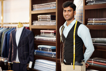 Portrait of handsome smiling Indian tailor with measuring tape around his neck standing in atelier...