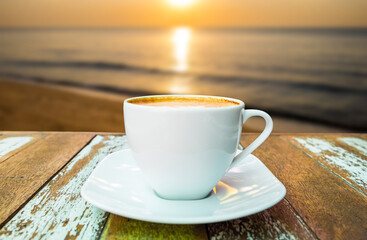 Front view of hot coffee cup on wooden table at sunset or sunrise on the beach background.
