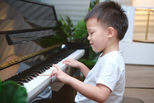 Cute happy smiling little Asian kid boy playing piano in living room at home, Preschool child having fun with learning to play music instrument