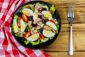 Tasty tuna salad with lettuce, black olives, eggs and fresh vegetables on wooden table. Top view