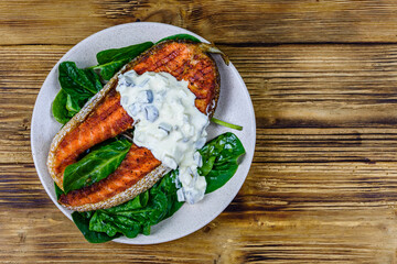 Plate with roasted salmon steak, tartar sauce and spinach leaves on a wooden table. Top view