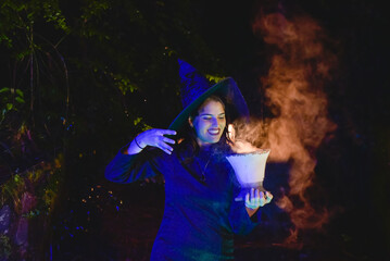 Halloween witch young girl wicca wearing black costume witches holding a old clay pot  for a spells and magic lights in a dark forest  