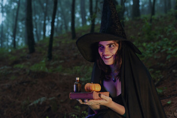 Halloween witch young girl wicca wearing black costume witches holding a old book for a spells and pumpkin in a dark forest  with magic lights