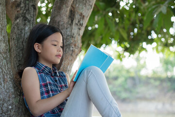  little asian girl reading a book under big tree. children and science. blurred background. learning the imagination and dreams of rural child. studying at home.