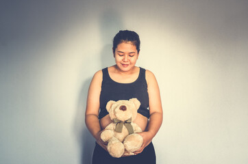 Happy pregnant Asian woman holding cute brown teddy bear over her belly on wall background at home....