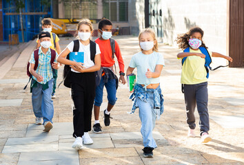 Multiethnic group of tweenagers in medical masks walking outside school building on autumn day, going to lessons. Concept of necessary .precautions in COVID pandemic..