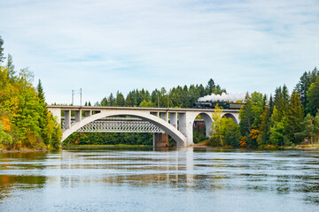 Fototapeta premium Kouvola, Finland - 18 September 2020: Autumn landscape of bridge with moving old steam passenger train Ukko-Pekka and Kymijoki river waters in Finland, Kouvola, Koria