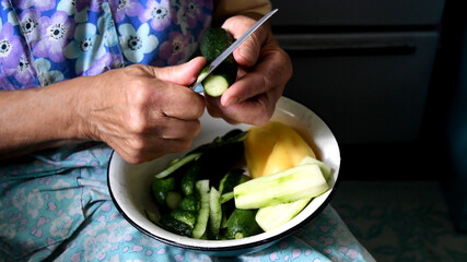 Female mature hands peel and cut cucumber into bowl of fresh vegetables