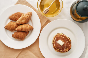 Top view of stack of sweet tasty pancakes, croissants, honey in a bowl with dipper and a teapot, Breakfast served on the table