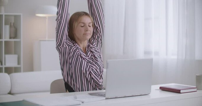 student woman is learning online with laptop and copybook at home, stretching tired muscles of back and neck