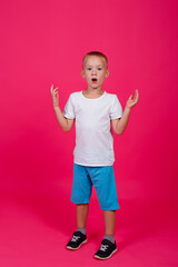 A very cute young surprised boy in full height looks at the camera, holding his hands up, isolated on a pink background.